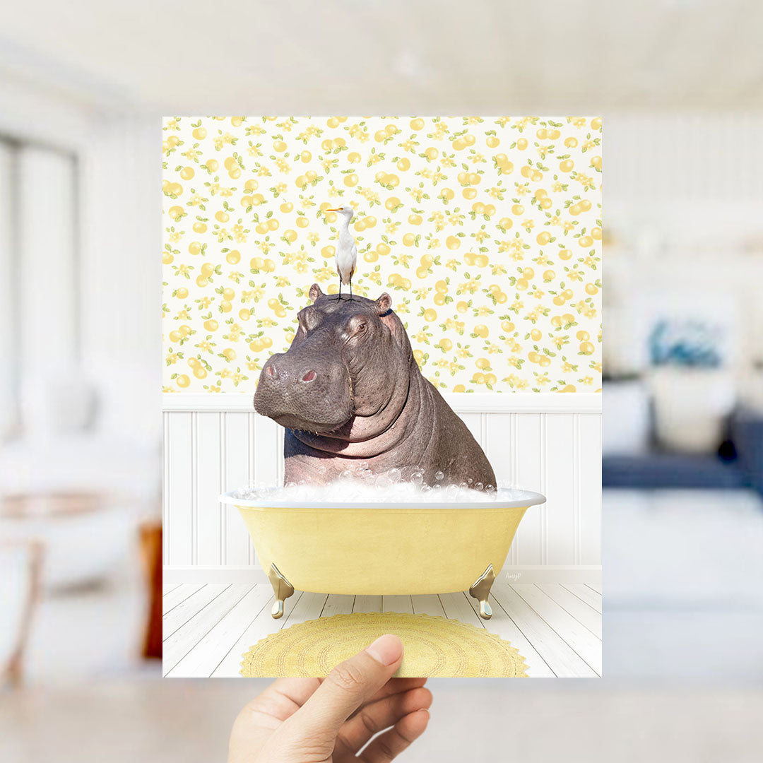 A hand holds up a framed image depicting a hippopotamus sitting in a yellow bathtub, surrounded by a yellow and white floral pattern.