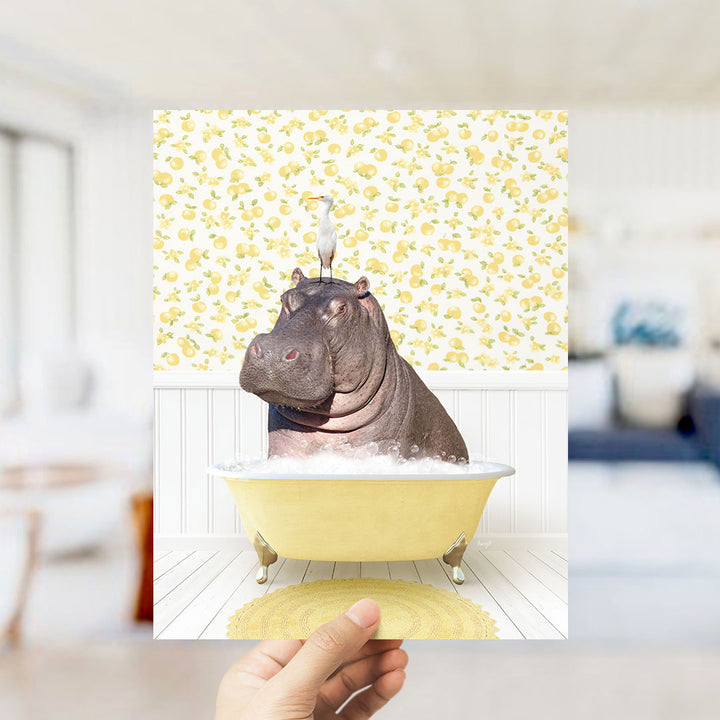 A hand holds up a framed image depicting a hippopotamus sitting in a yellow bathtub, surrounded by a yellow and white floral pattern.