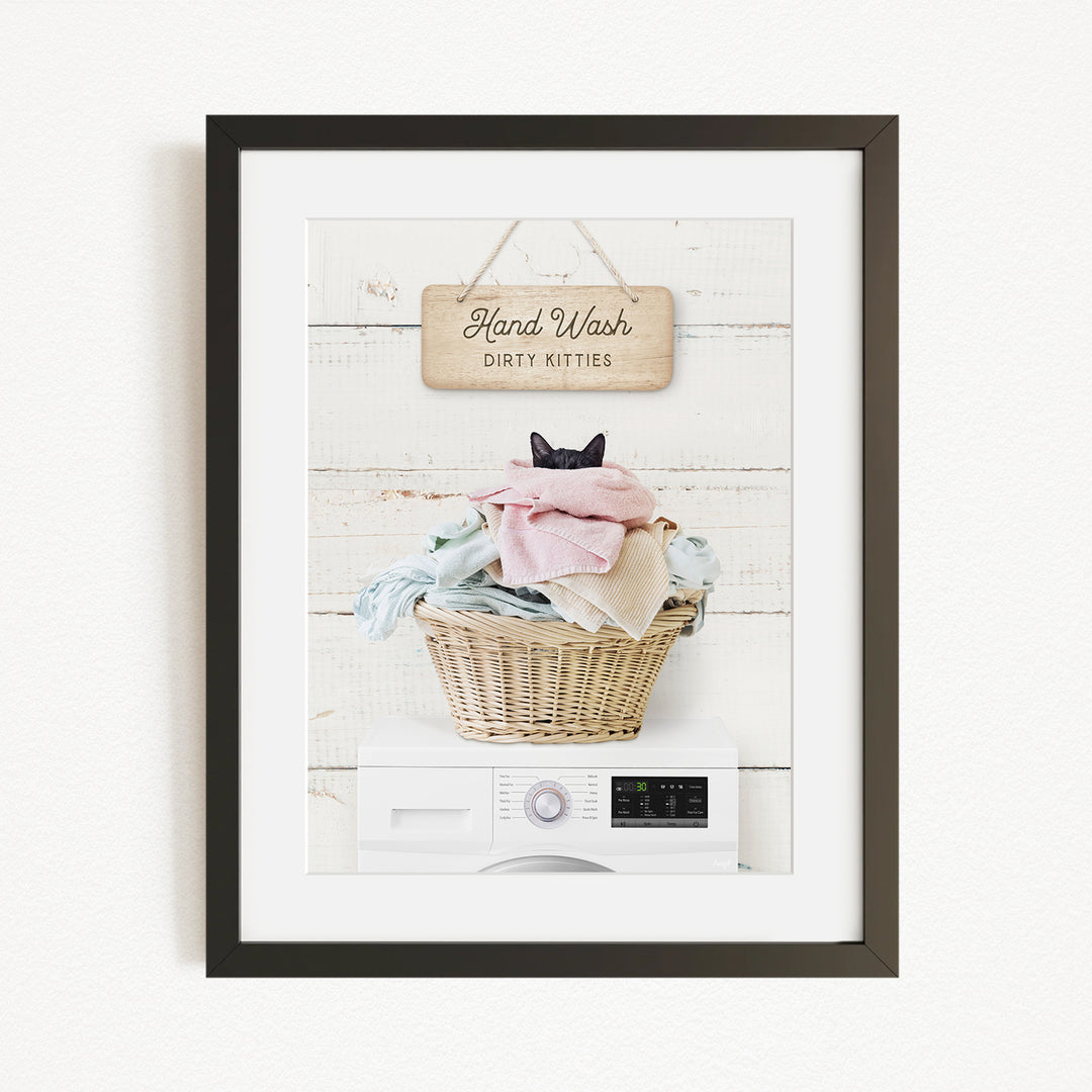 A framed image depicting a cat sitting in a basket filled with laundry, with a sign above that reads "Hand Wash Dirty Kittens".