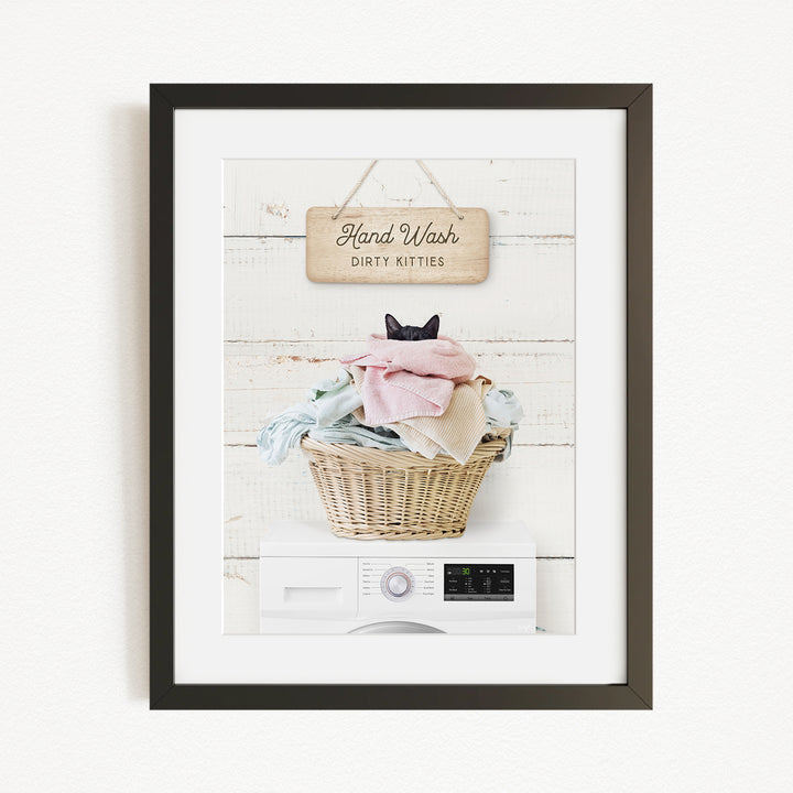 A framed image depicting a cat sitting in a basket filled with laundry, with a sign above that reads "Hand Wash Dirty Kittens".