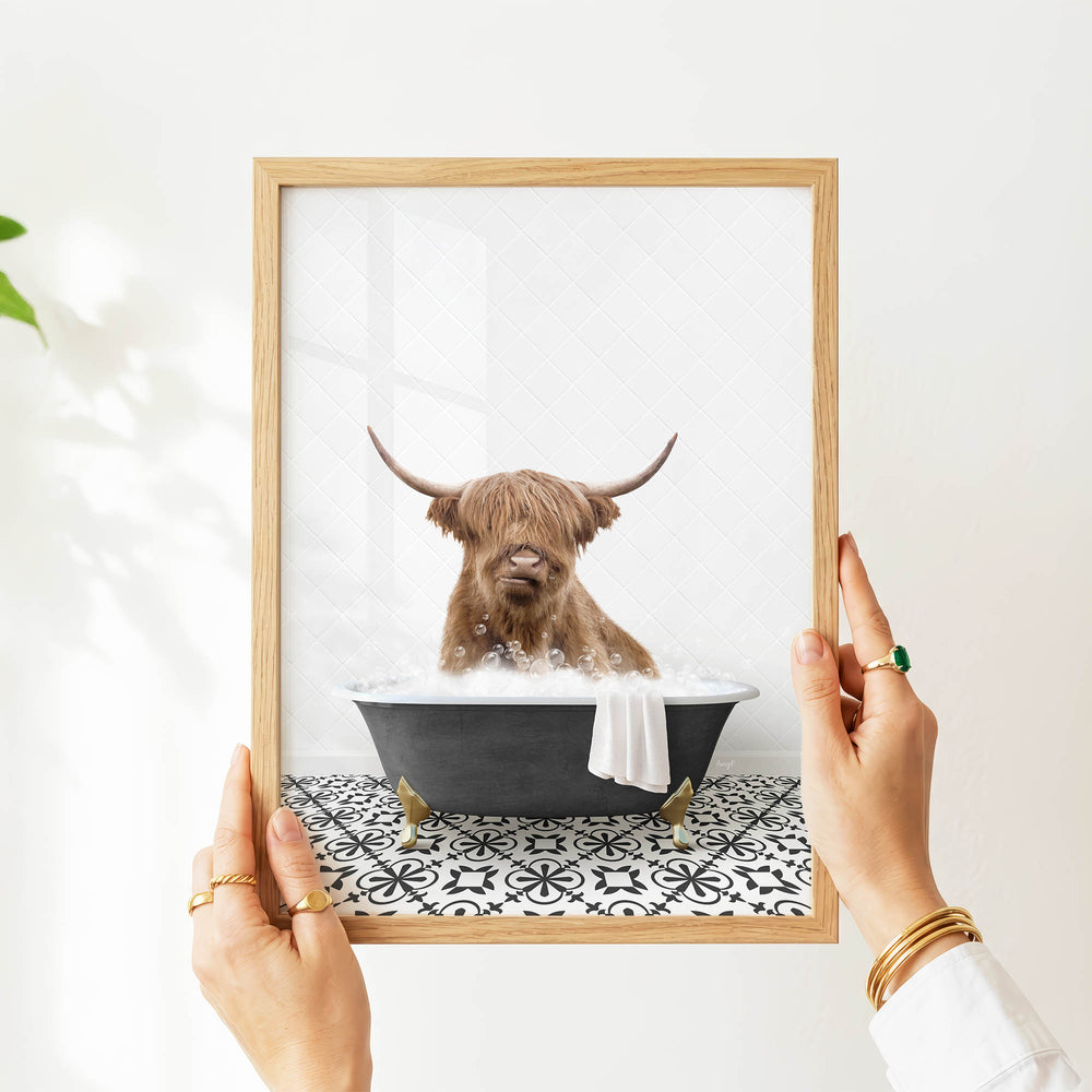 A person's hands are holding a framed picture of a Highland cow sitting in a bathtub filled with water and bubbles.