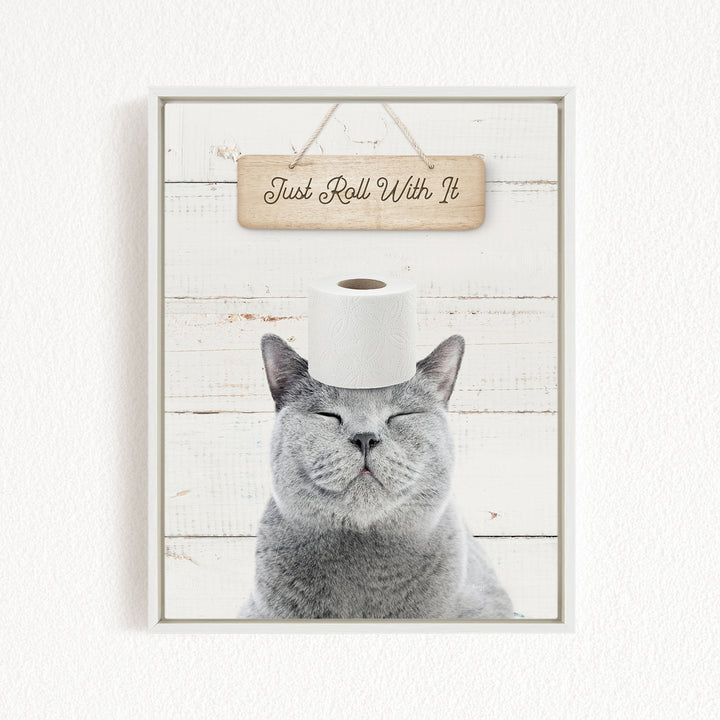 A gray cat with its eyes closed and a roll of toilet paper on its head, with a wooden sign above that says "Just Roll With It".