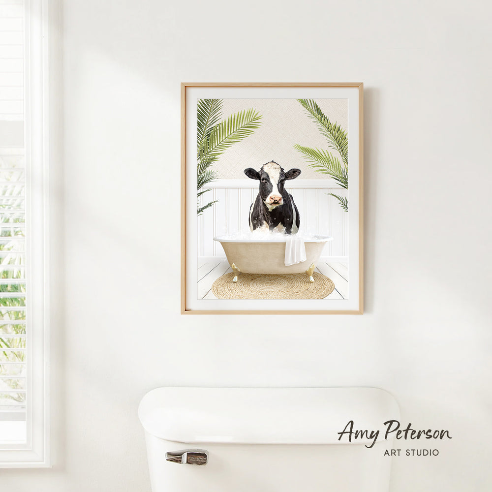 A framed image of a cow sitting in a bathtub, surrounded by palm leaves, is hanging on a white wall in a bathroom.
