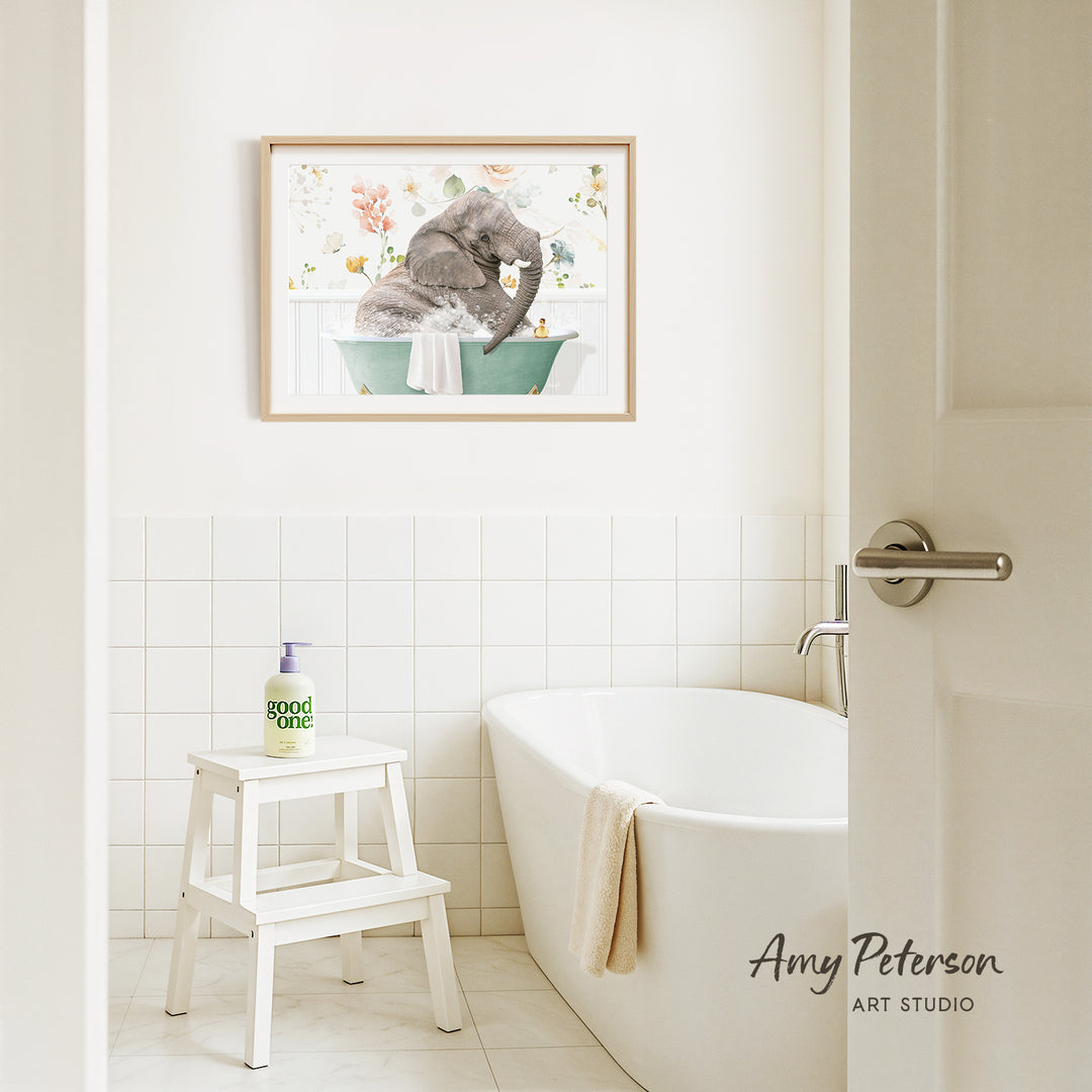A framed painting of an elephant sitting in a bathtub is hanging on a white tiled wall above a white bathtub in a bathroom.