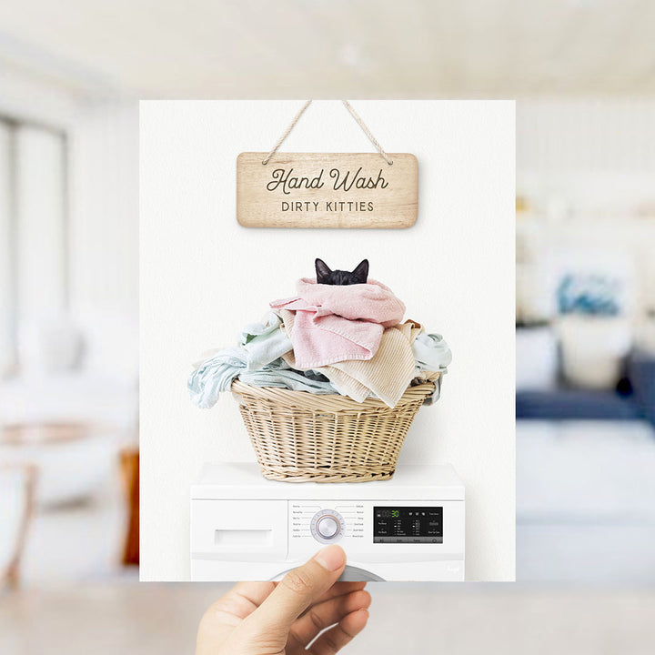 A hand holding a sign that says "Hand Wash Dirty Kitties" above a washer with a basket of clothes inside.