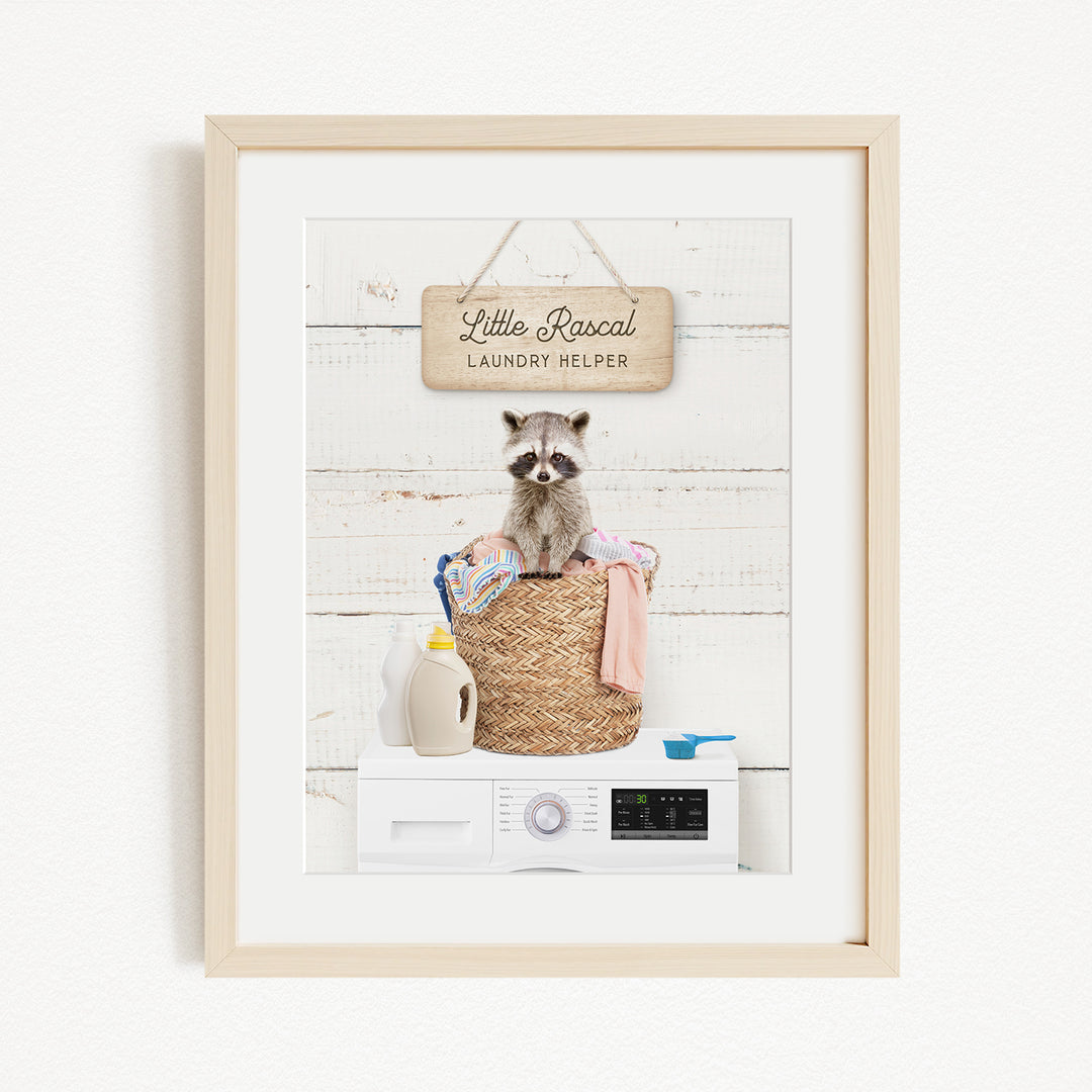 A framed image depicting a raccoon sitting in a laundry basket, surrounded by various household items such as a washing machine, detergent, and a sign that reads "Little Racal Laundry Helper".
