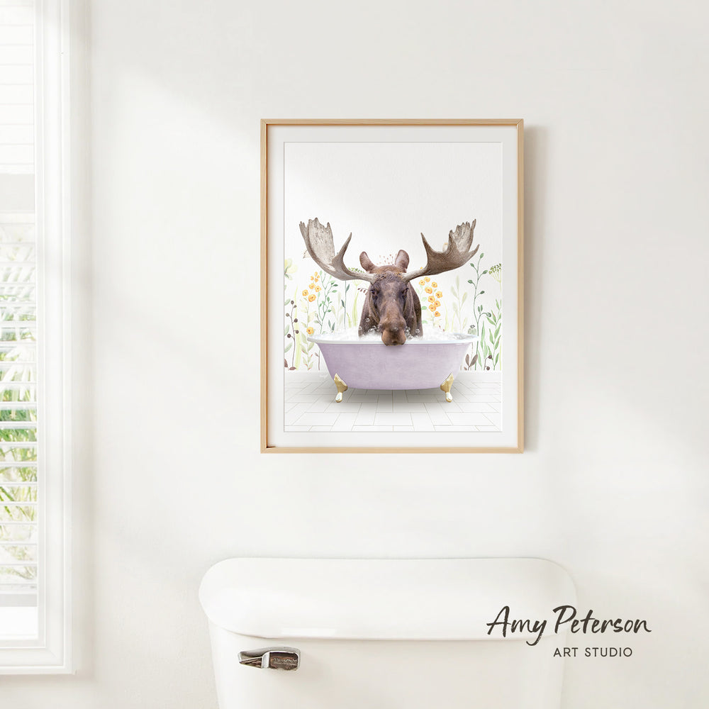 A framed print of a moose sitting in a bathtub is hanging on a white wall.