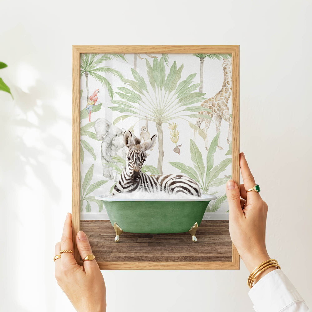 A person's hands are holding a framed picture of a zebra sitting in a green bathtub, surrounded by tropical foliage.