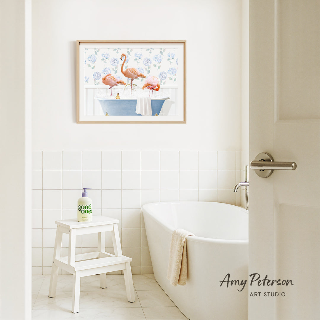 A framed painting of two pink flamingos sitting in a blue bathtub is hanging on a white tiled wall above a white bathtub in a bathroom.