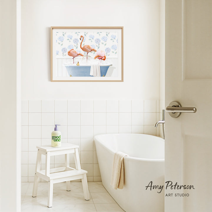 A framed painting of two pink flamingos sitting in a blue bathtub is hanging on a white tiled wall above a white bathtub in a bathroom.
