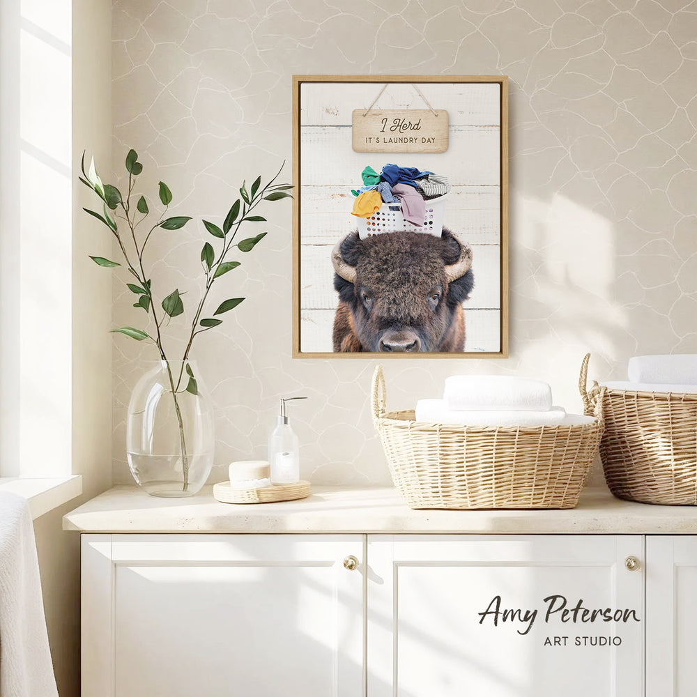 a framed picture of a bison wearing a hat with a sign that says "I herd it's laundry day" hanging on a wall above a white cabinet.