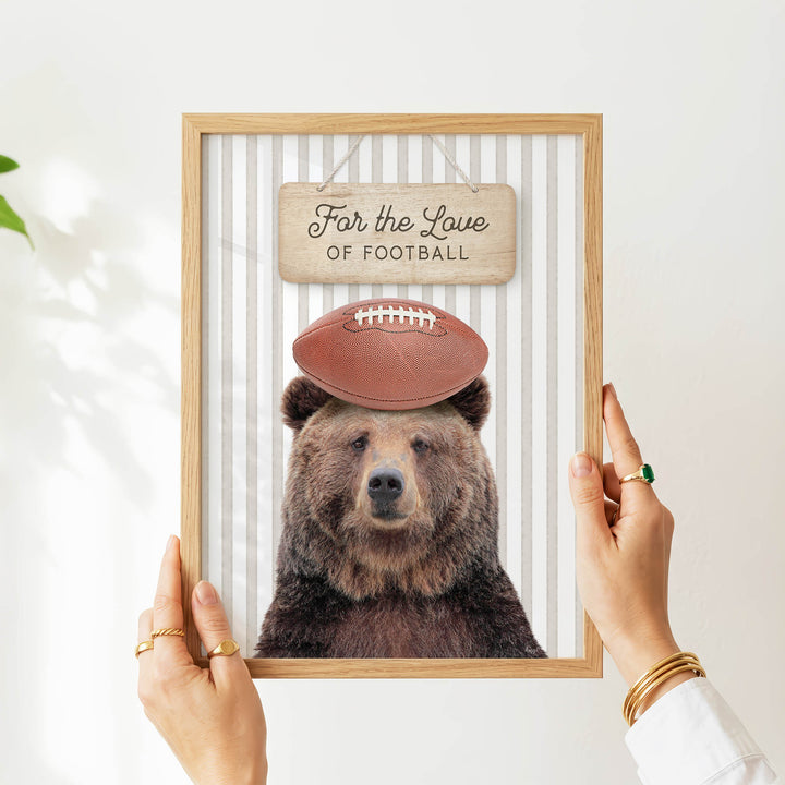 A person's hands are holding a framed picture of a bear wearing a football helmet, with a sign above the bear that says "For the Love of Football".