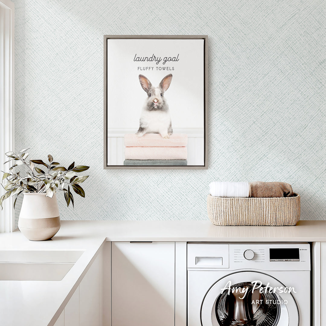 a laundry room with a framed poster of a rabbit sitting on a stack of fluffy towels, a potted plant, and a basket of folded towels on a counter.