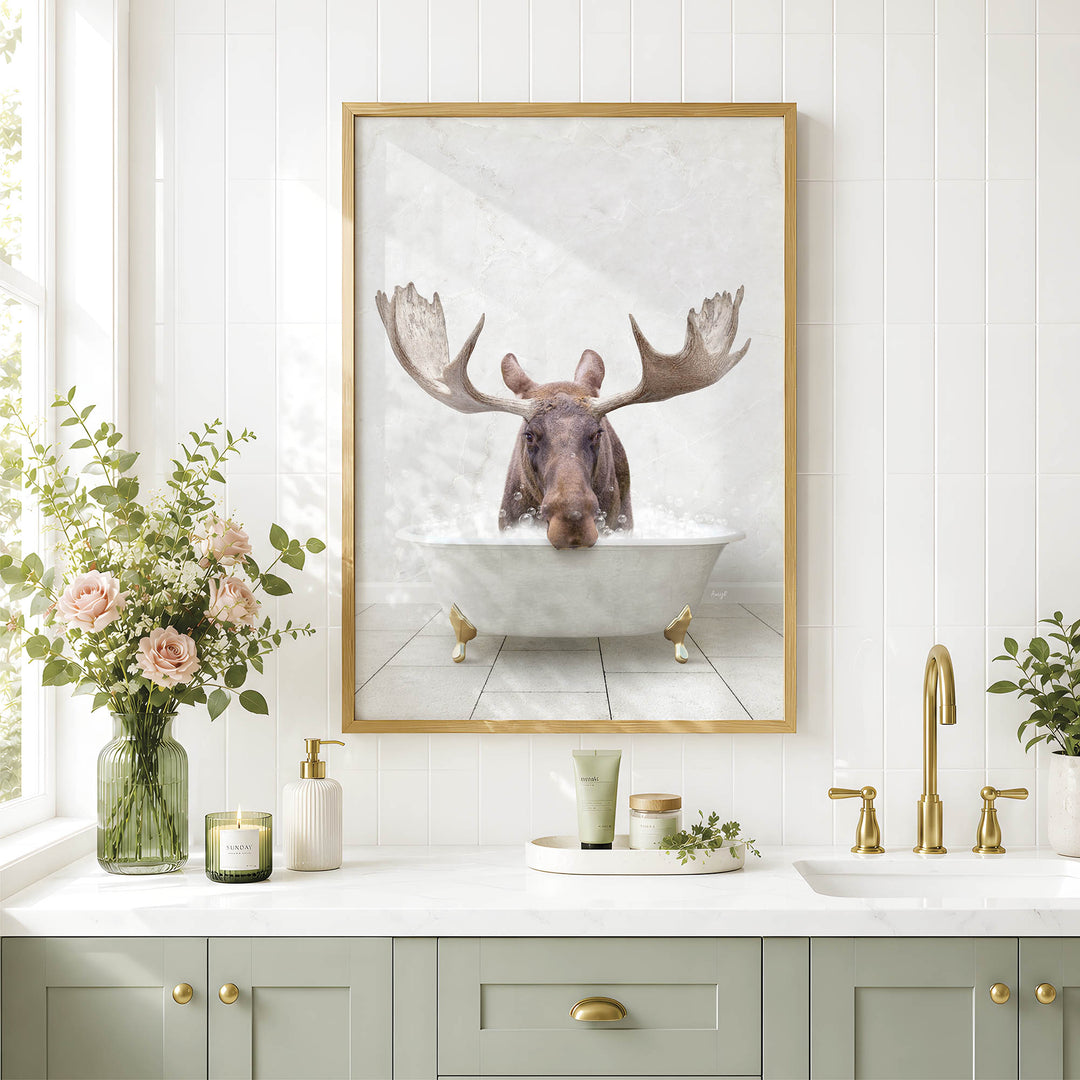 a framed picture of a moose in a bathtub, surrounded by a vase of pink flowers, a candle, and a tray of candles on a white countertop.