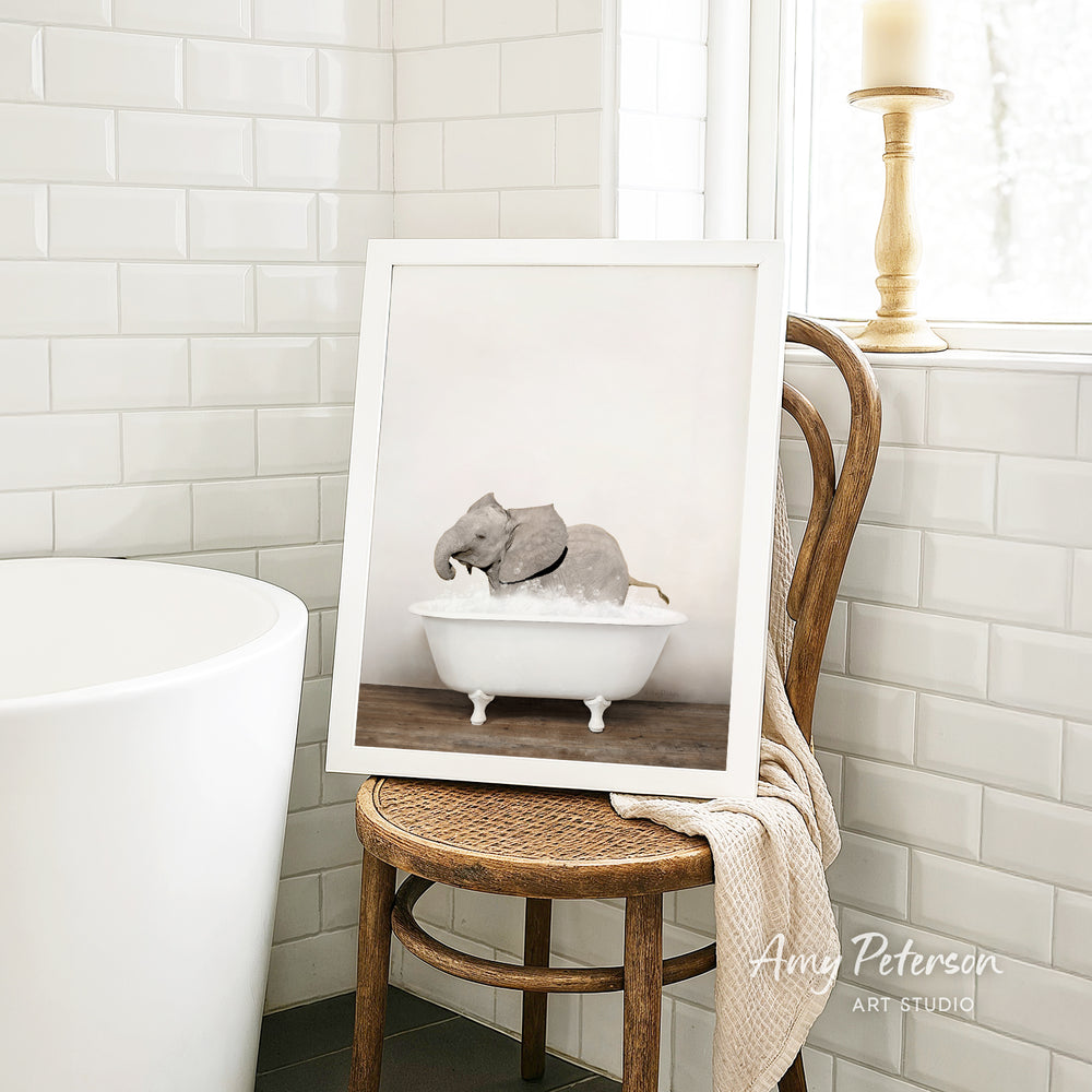A framed photograph of a baby elephant sitting in a bathtub is displayed on a wooden chair in a bathroom setting.