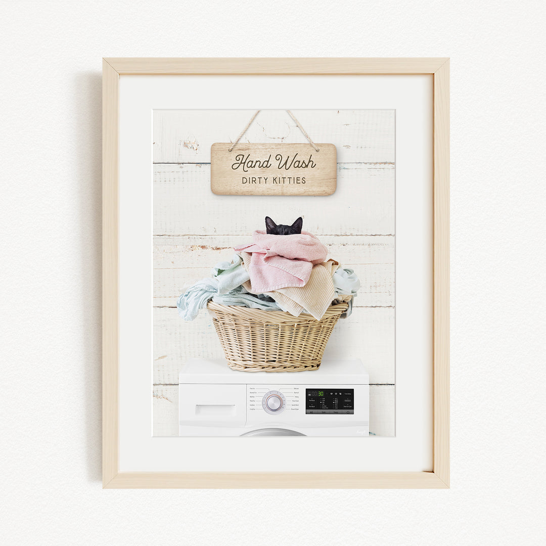A framed image depicting a cat sitting in a basket filled with laundry, with a sign above that reads "Hand Wash Dirty Kittens".