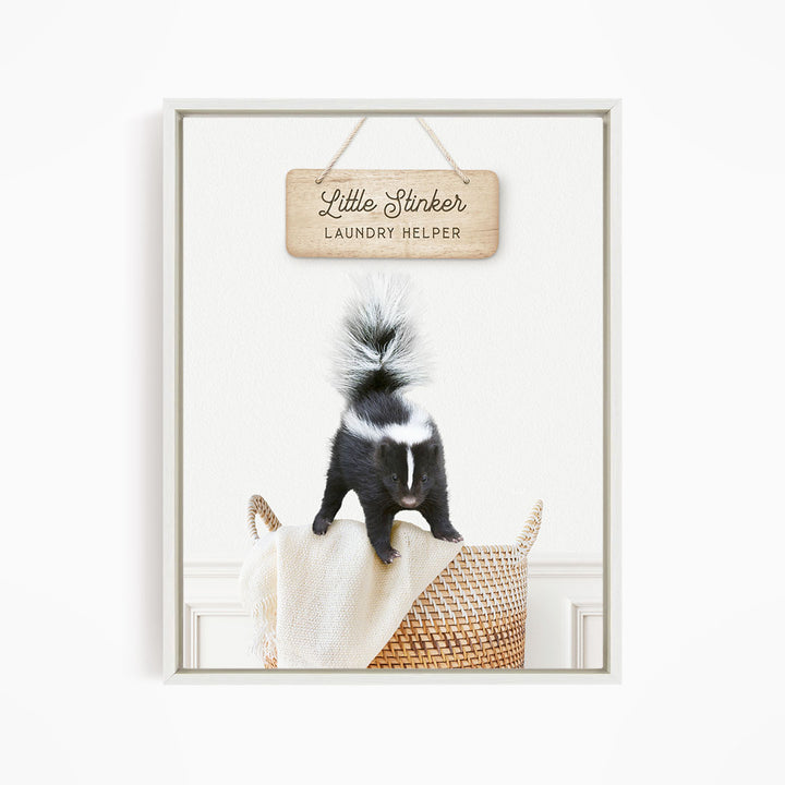 A black and white striped skunk standing on a woven basket, with a wooden sign above that reads "Little Stinker Laundry Helper".