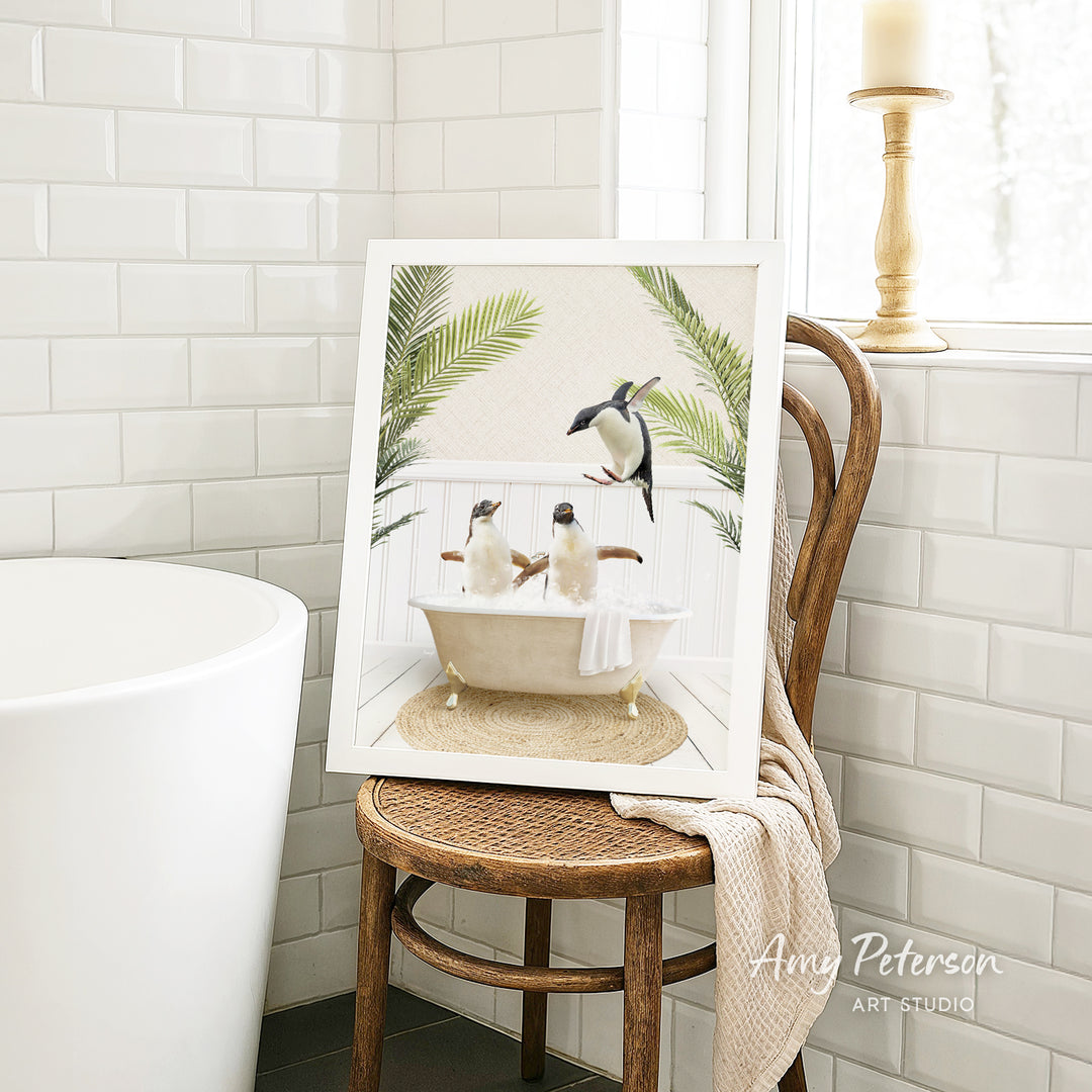 A framed image of two penguins sitting in a bathtub, surrounded by palm leaves, is displayed on a wooden chair in a bathroom setting.