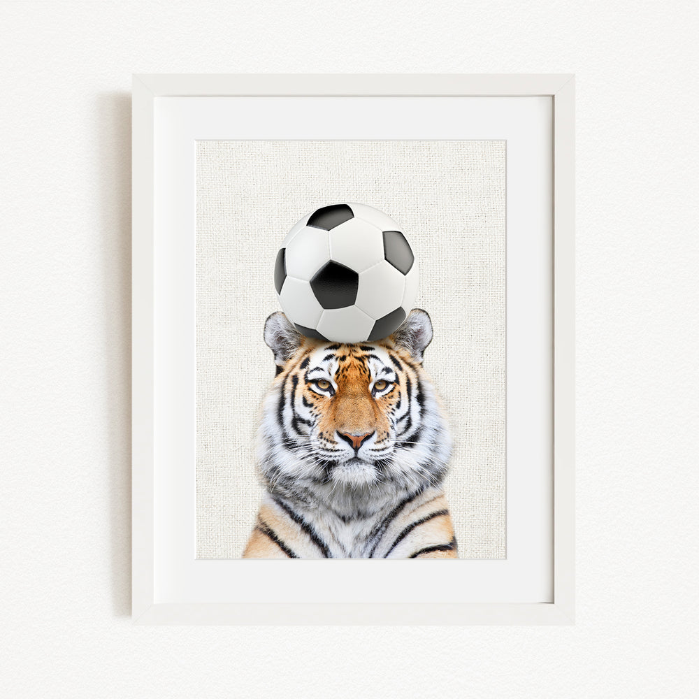 A tiger is sitting in front of a soccer ball, with the ball balanced on its head.