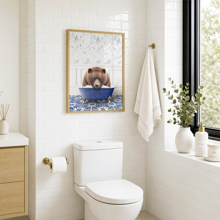 A bathroom with a white toilet, a framed picture of a bear sitting in a blue bathtub, and a window with greenery outside.