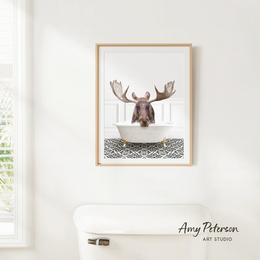 A framed image of a moose sitting in a bathtub, with its antlers spread out, is hanging on a white wall.