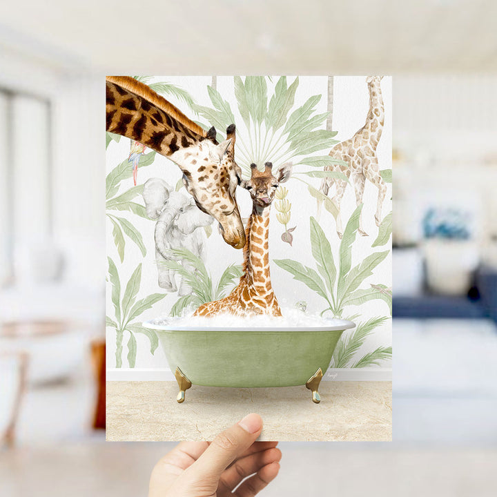 A hand holds up a framed image depicting a giraffe and a baby giraffe in a bathtub surrounded by tropical foliage.