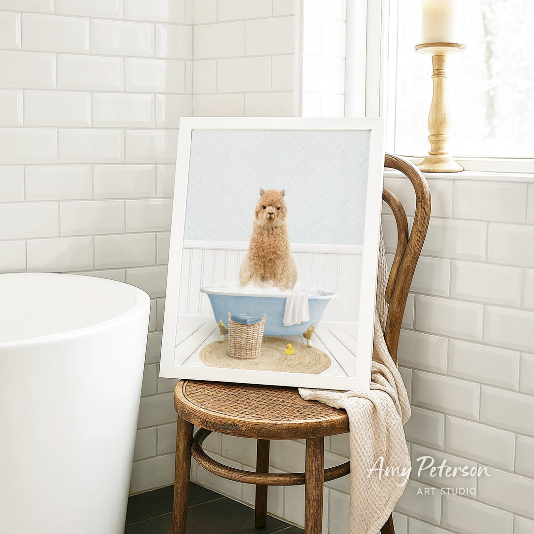 A framed image of a small dog sitting in a bathtub, surrounded by a blue tub, a basket, and a towel, is displayed on a wooden chair in a bathroom setting.