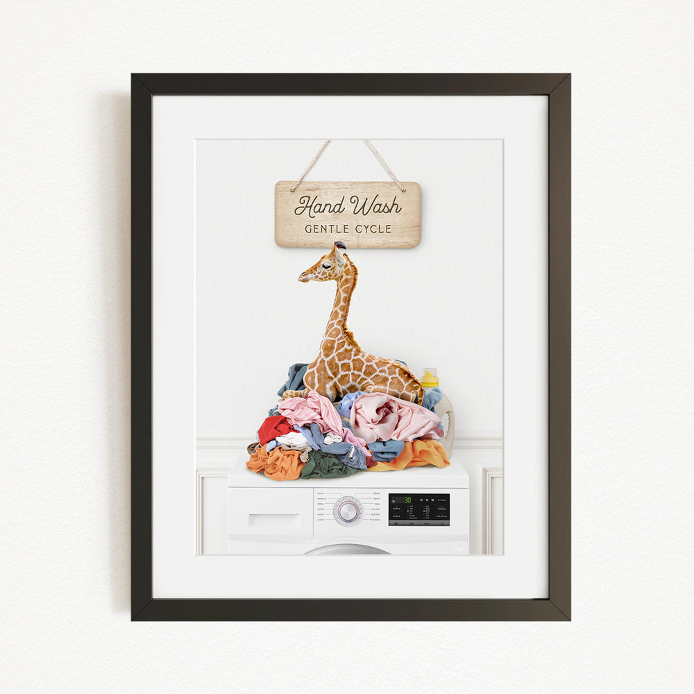 A framed image depicting a giraffe sitting on a pile of laundry, with a sign above that reads "Hand Wash Gentle Cycle".