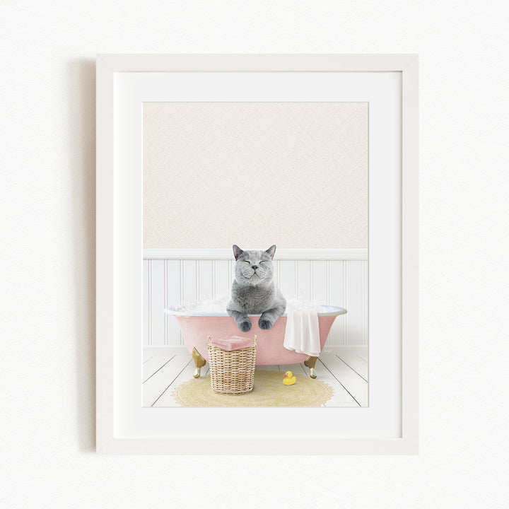 A gray cat sitting in a pink bathtub, with a yellow rubber duck nearby and a basket of bath toys on the floor.