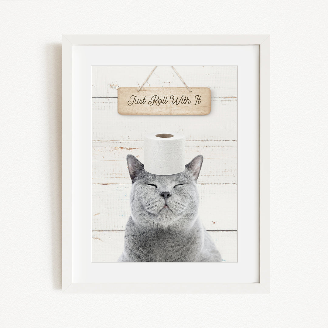 A gray cat with its eyes closed, wearing a white toilet paper roll on its head, with a wooden sign above that says "Just Roll With It".