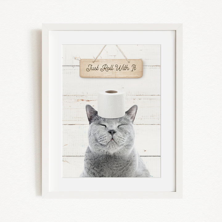 A gray cat with its eyes closed, wearing a white toilet paper roll on its head, with a wooden sign above that says "Just Roll With It".