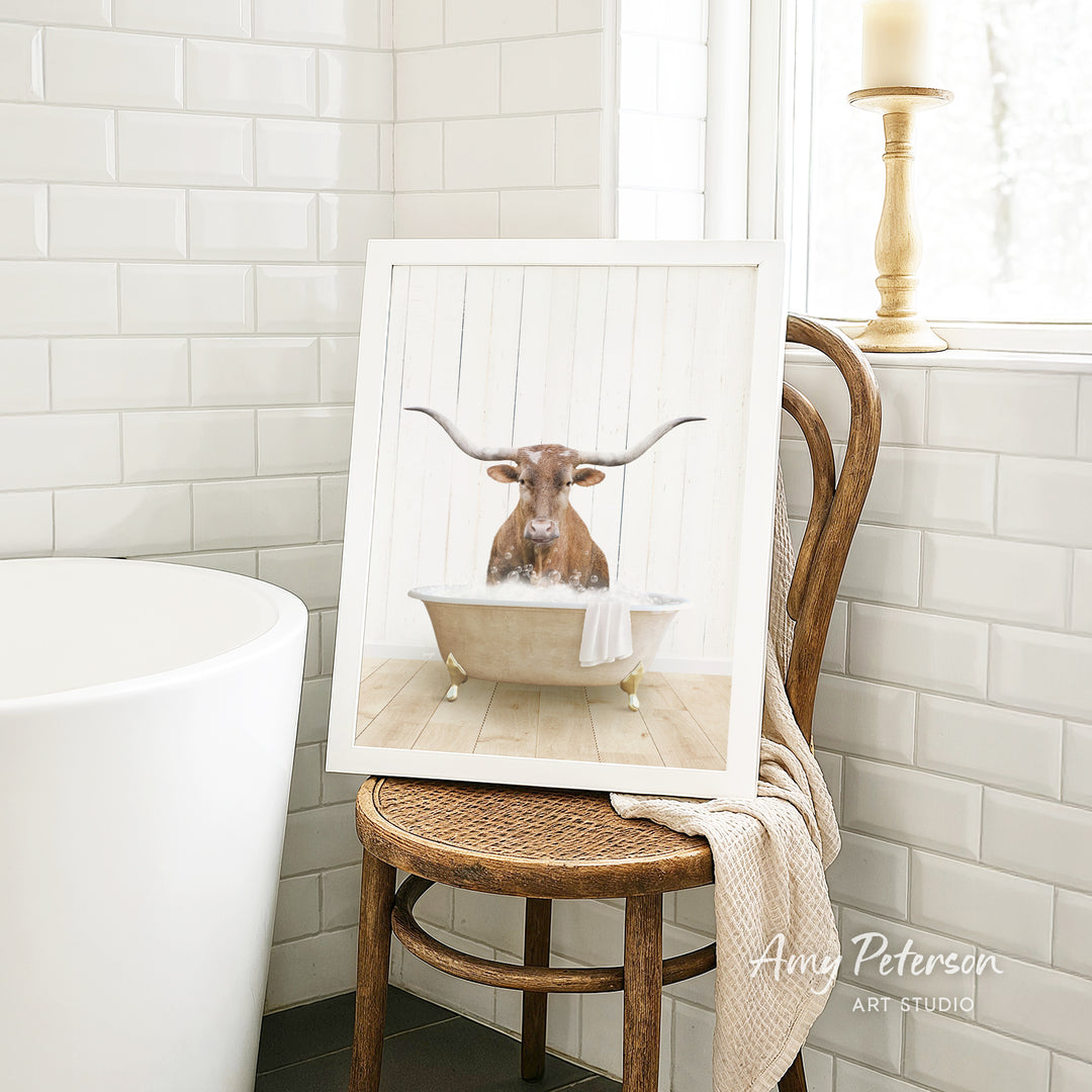 A framed image of a bull with long horns sitting in a bathtub, surrounded by a towel, is displayed on a wooden chair in a bathroom setting.