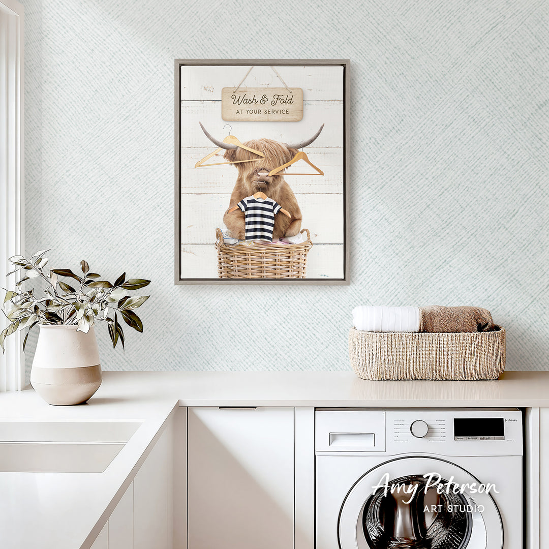 a framed picture of a cow sitting in a basket, with a sign above it that says "Wash & Fold at Your Service". The picture is displayed on a wall above a white countertop, which has a potted plant and a basket of towels on it. There is also a washing machine visible in the background.