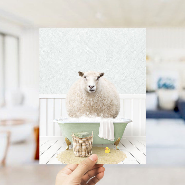 A hand holds up a framed image of a sheep sitting in a bathtub filled with bubbles, surrounded by a basket of bath toys and a yellow rubber duck.