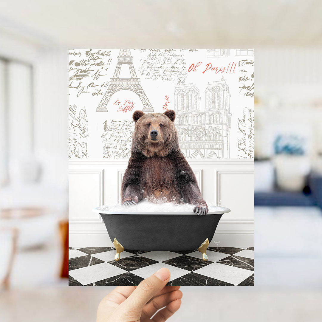 A hand holds up a framed image depicting a brown bear sitting in a bathtub, with the Eiffel Tower and Parisian buildings visible in the background.