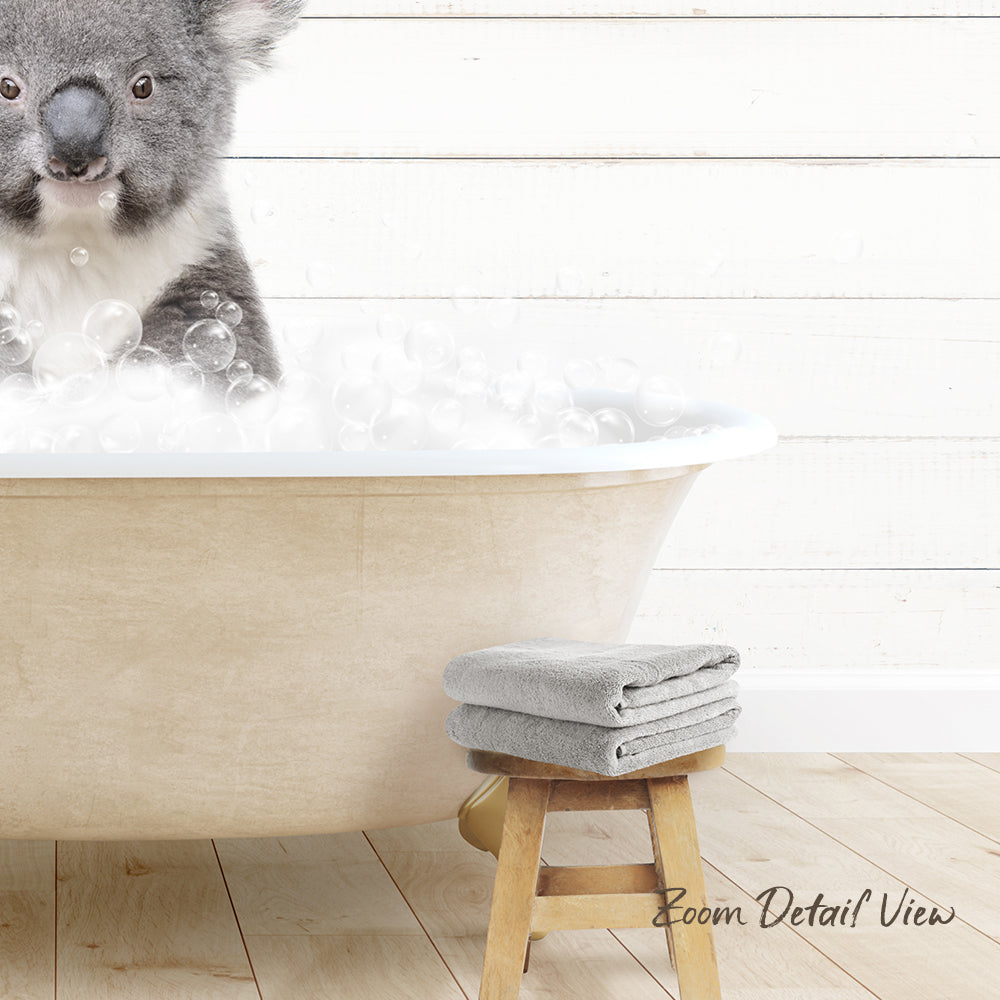 A koala sitting in a bathtub filled with bubbles, with a towel and stool nearby.