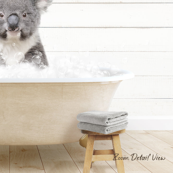 A koala sitting in a bathtub filled with bubbles, with a towel and stool nearby.
