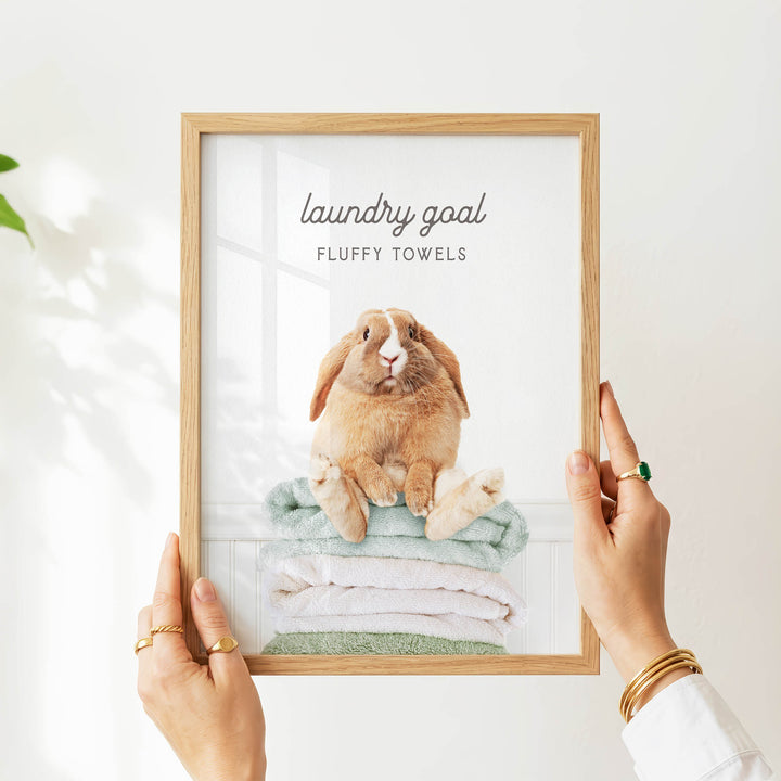 A person is holding up a framed picture of a rabbit sitting on a stack of fluffy towels, with the text "laundry goal" and "fluffy towels" written above the image.