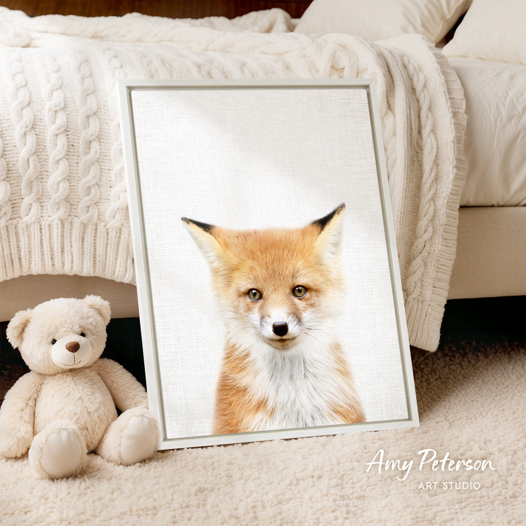 A framed print of a fox's face is displayed on a white bedspread, accompanied by a teddy bear on the floor.