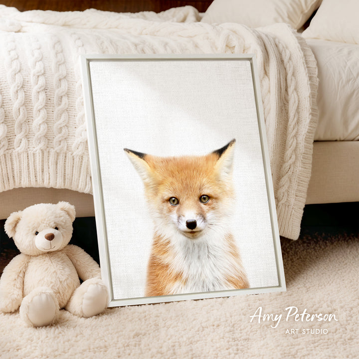 A framed print of a fox's face is displayed on a white bedspread, accompanied by a teddy bear on the floor.