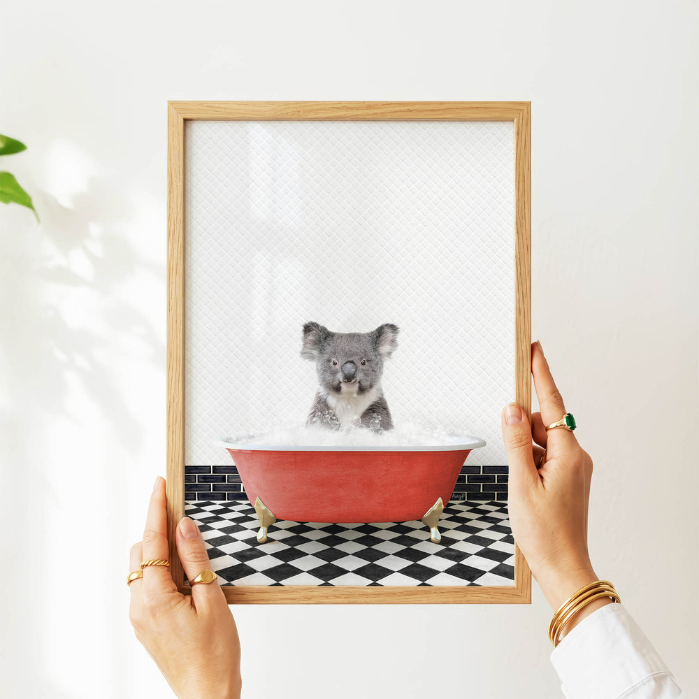 A person's hands are holding a framed picture of a koala bear sitting in a red bathtub filled with water.