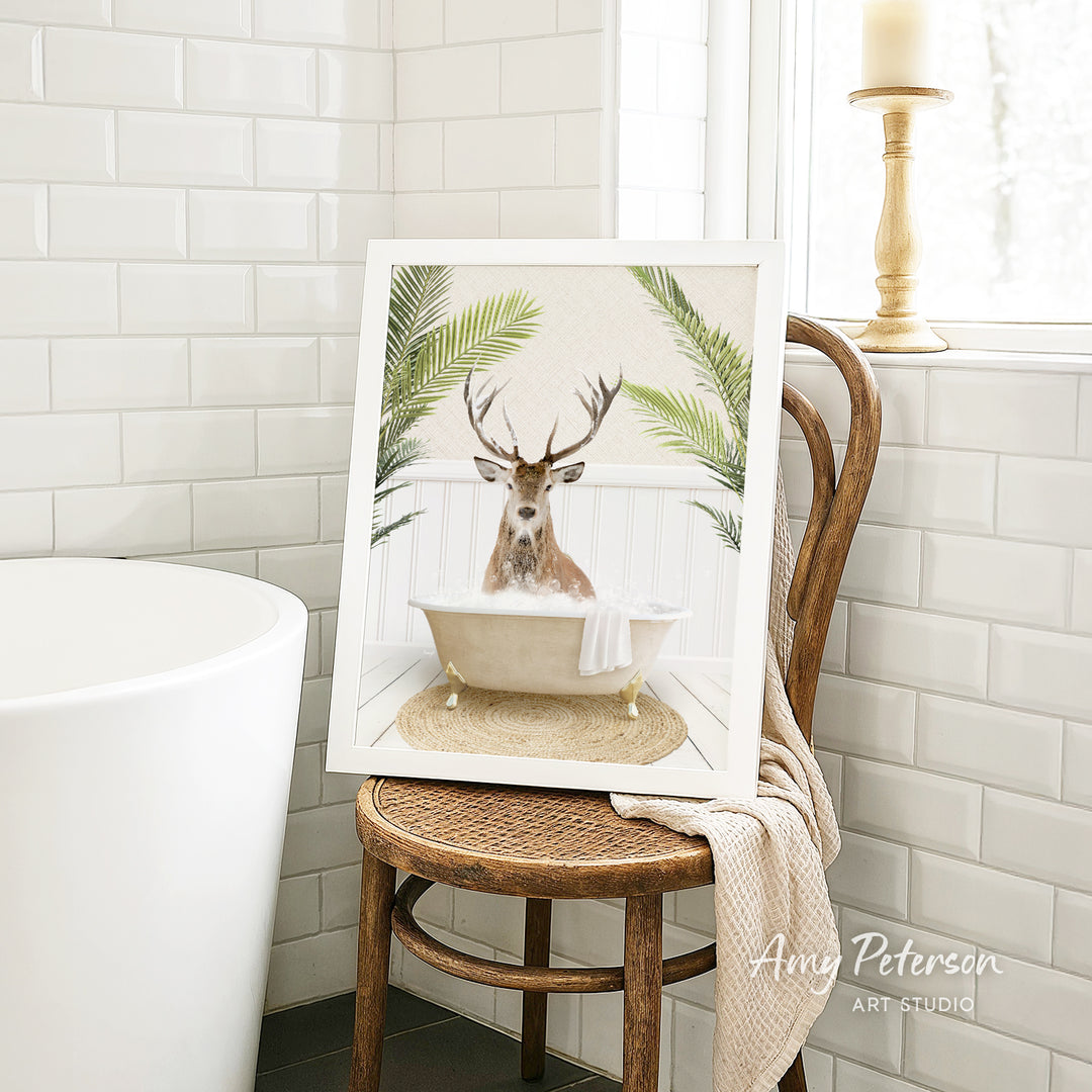 A framed image of a deer sitting in a bathtub, surrounded by palm leaves, is displayed on a wooden chair in a bathroom setting.