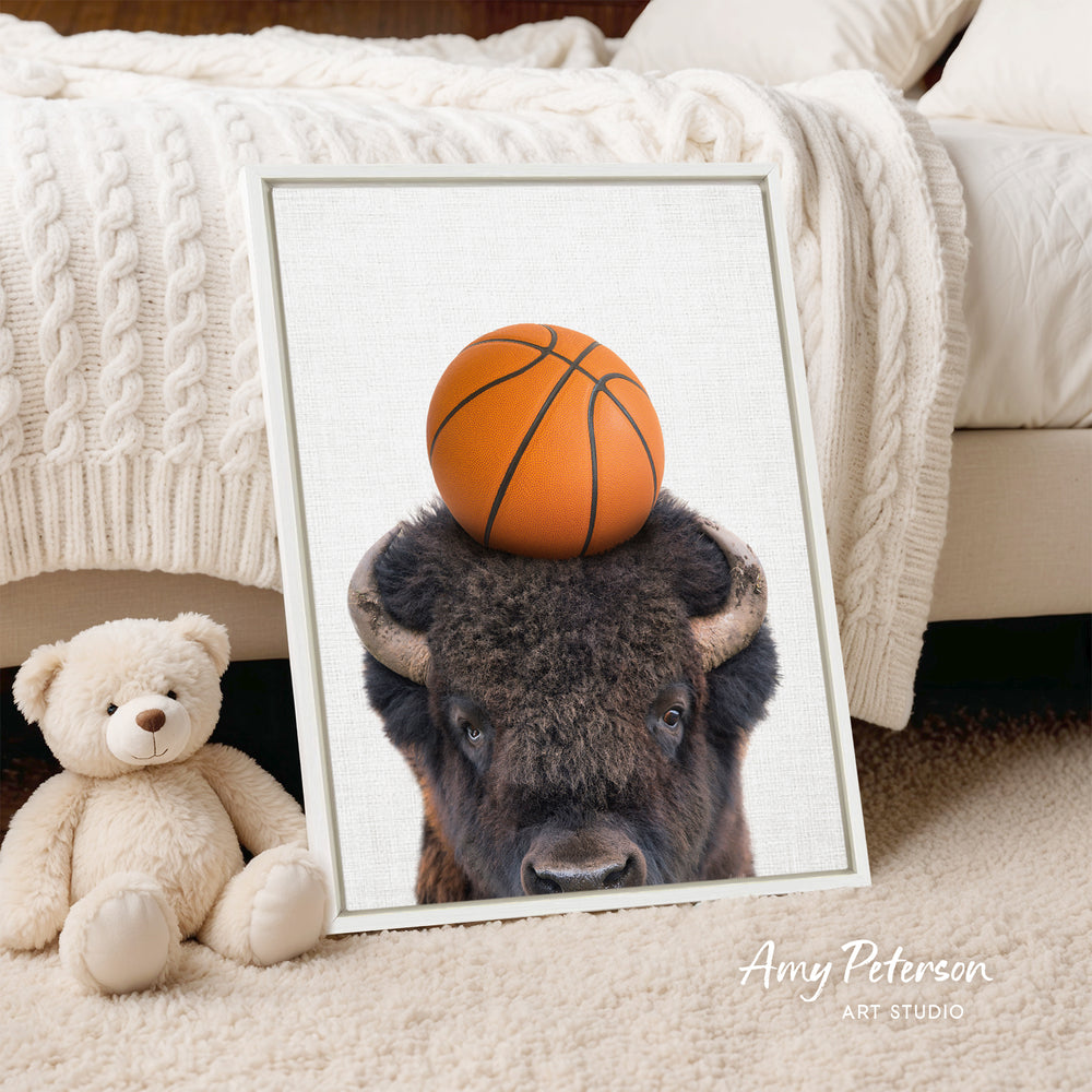 A framed image of a buffalo with a basketball on its head, placed on a bed next to a stuffed teddy bear.