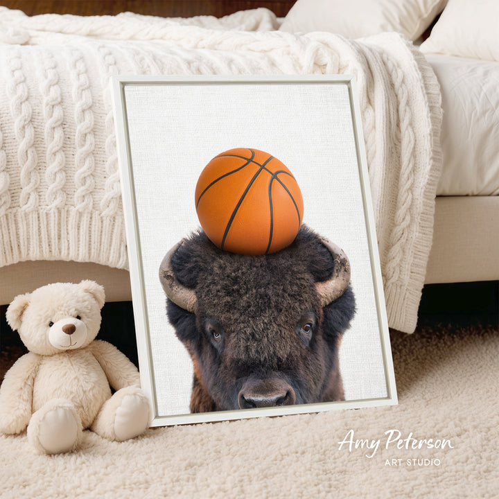 A framed image of a buffalo with a basketball on its head, placed on a bed next to a stuffed teddy bear.