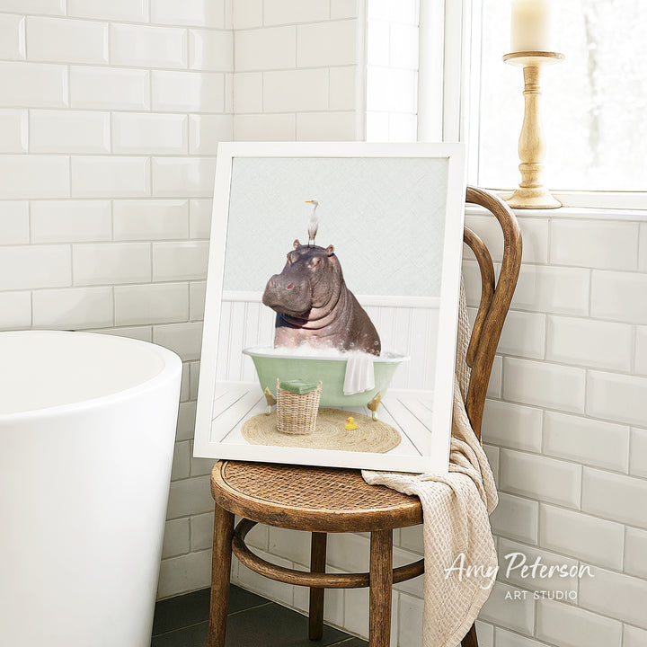 A framed image of a hippopotamus sitting in a bathtub, surrounded by a basket of bath toys, is displayed on a wooden chair in a bathroom setting.