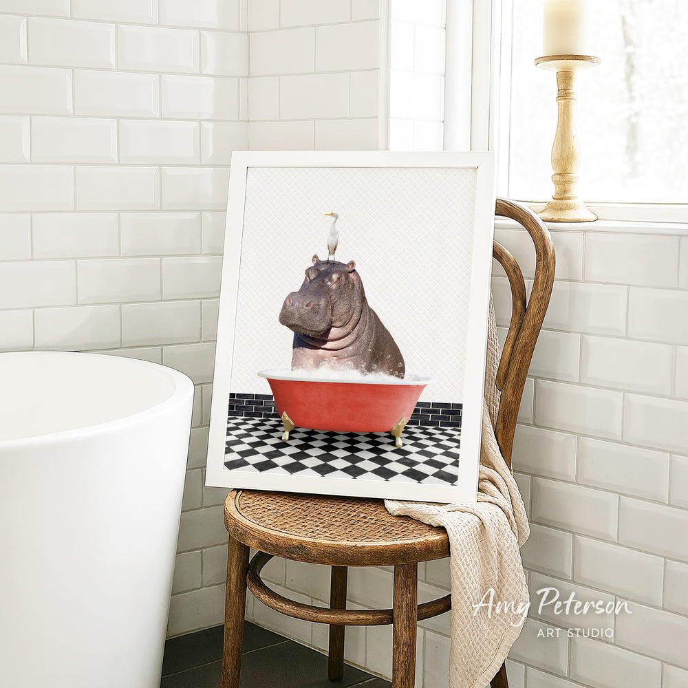 A framed image of a hippopotamus sitting in a red bathtub, with a bird perched on its head, is displayed on a wooden chair in a bathroom setting.