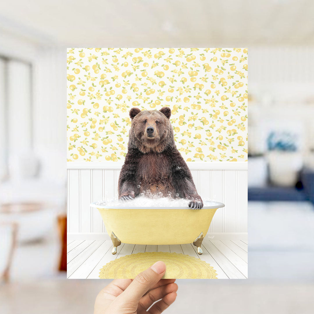 A hand holds up a framed image of a brown bear sitting in a yellow bathtub filled with bubbles, against a yellow and white floral wallpaper.
