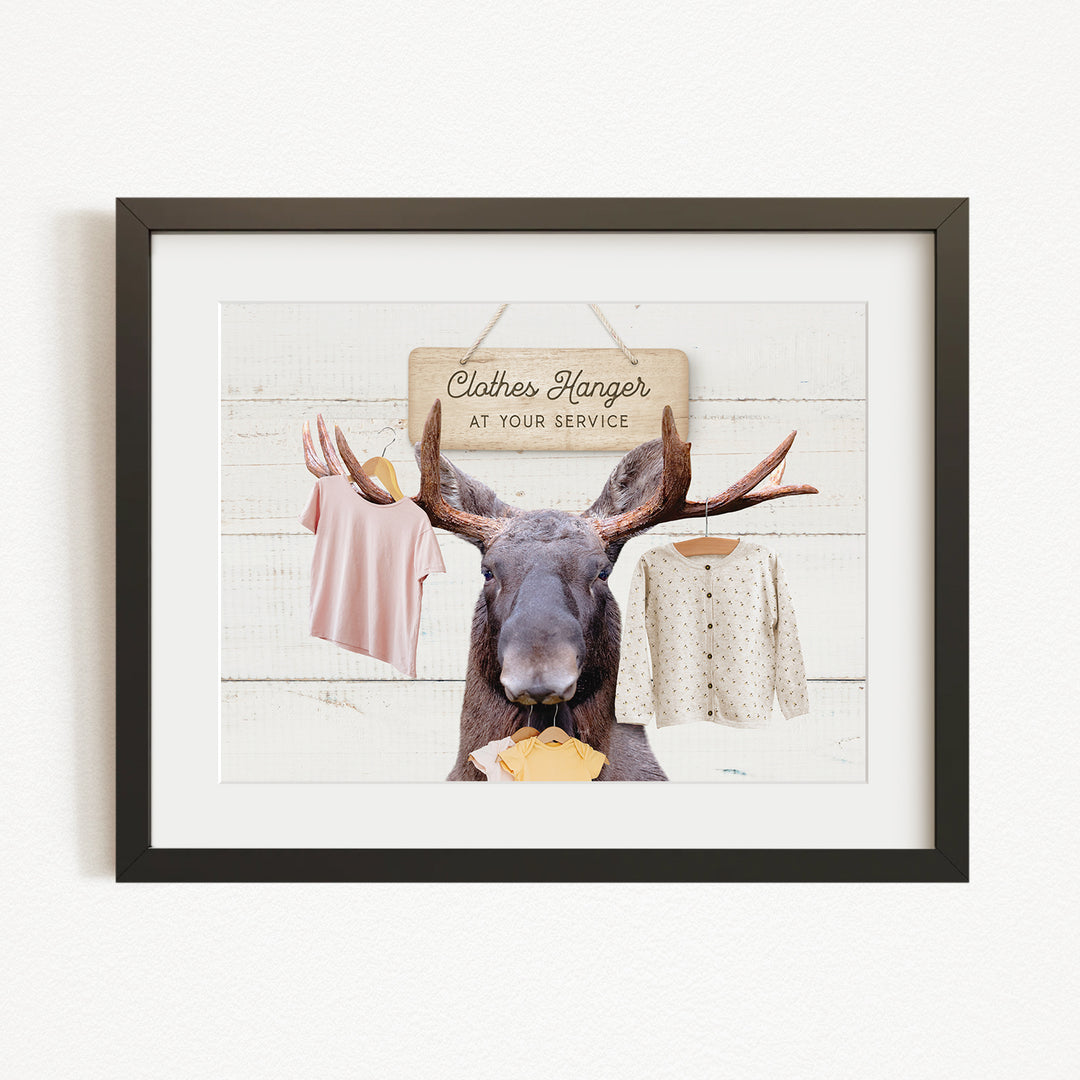 A framed image depicting a moose with antlers holding clothes in its mouth, with a sign that reads "Clothes Hanger at your service".