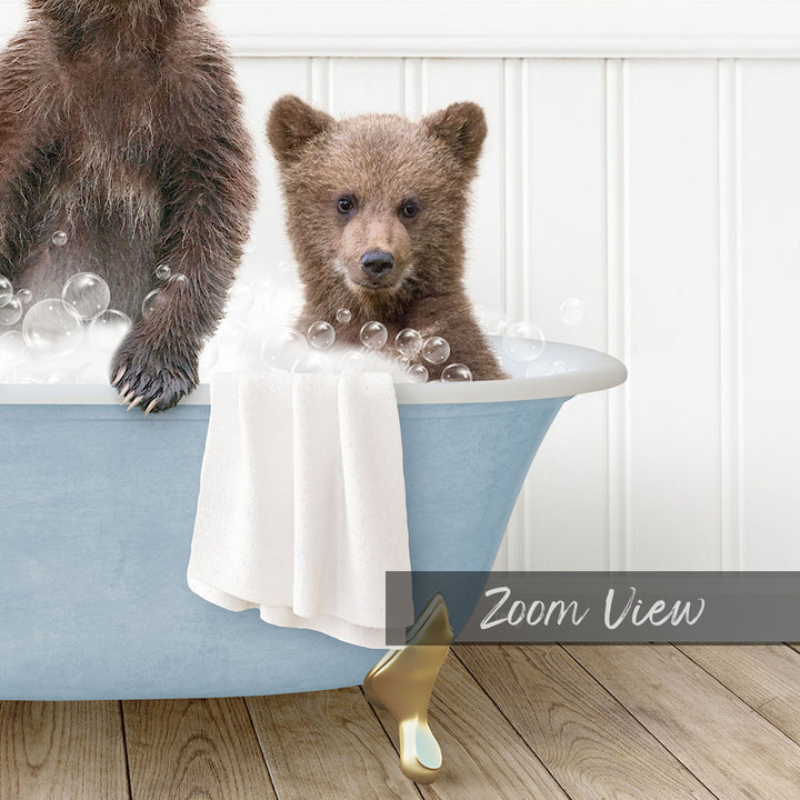 A baby bear sitting in a bathtub filled with bubbles, with a towel draped over the side of the tub.