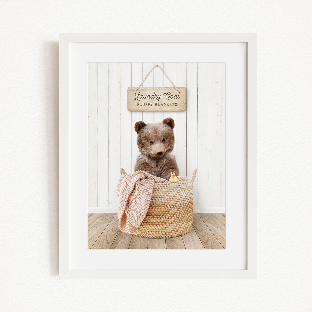 A framed image of a brown bear sitting in a woven basket, with a sign above the basket that reads "Laundry Goal Fluffy Blankets".